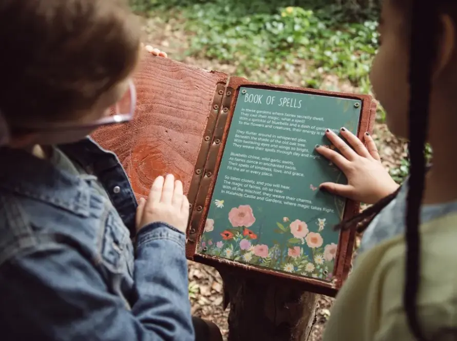 Two children outside in a forested area, dressed in casual clothes, are looking at an open book titled "Book of Spells." The book features intricate floral illustrations and spell descriptions. One child is reading while the other points to a spell on the page.