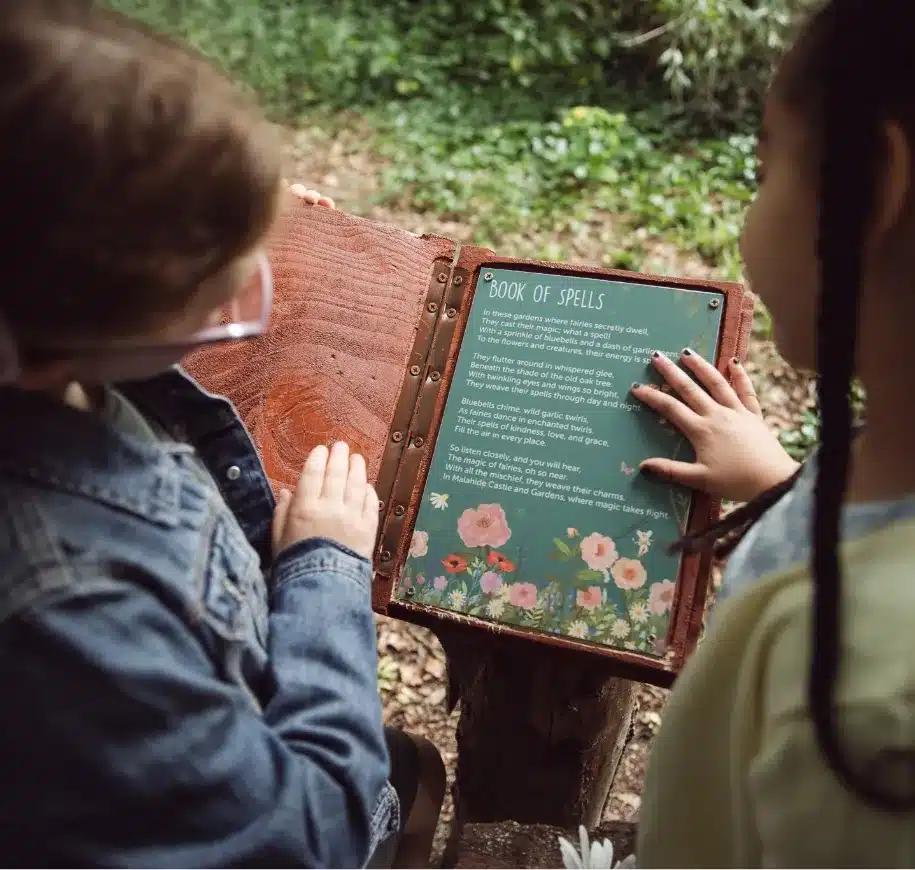 Two children outside in a forested area, dressed in casual clothes, are looking at an open book titled "Book of Spells." The book features intricate floral illustrations and spell descriptions. One child is reading while the other points to a spell on the page.