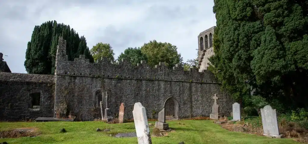 An ancient stone church with a crenelated wall, reminiscent of the historic charm at Malahide Castle, is surrounded by gravestones and tall trees. A grassy area lies in the foreground, while the church tower rises above the wall under overcast skies.