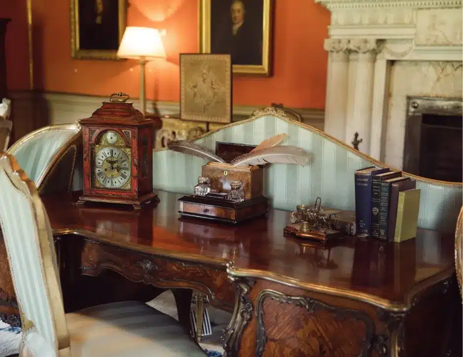 An antique wooden desk with ornate carvings is adorned with a vintage clock, quill pens, books, and a decorative lamp in a richly decorated room reminiscent of Malahide Castle. Paintings and a fireplace with a mantel grace the background.