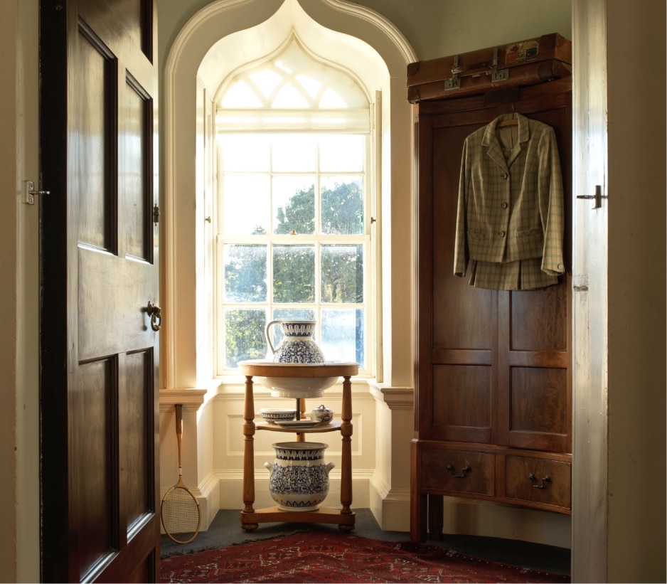 A sunlit room features a vintage wooden wardrobe with a plaid jacket hanging on it. In front is a small table with blue and white porcelain vases. A lattice window and a vintage tennis racket add to the decor. A red patterned rug covers the floor.