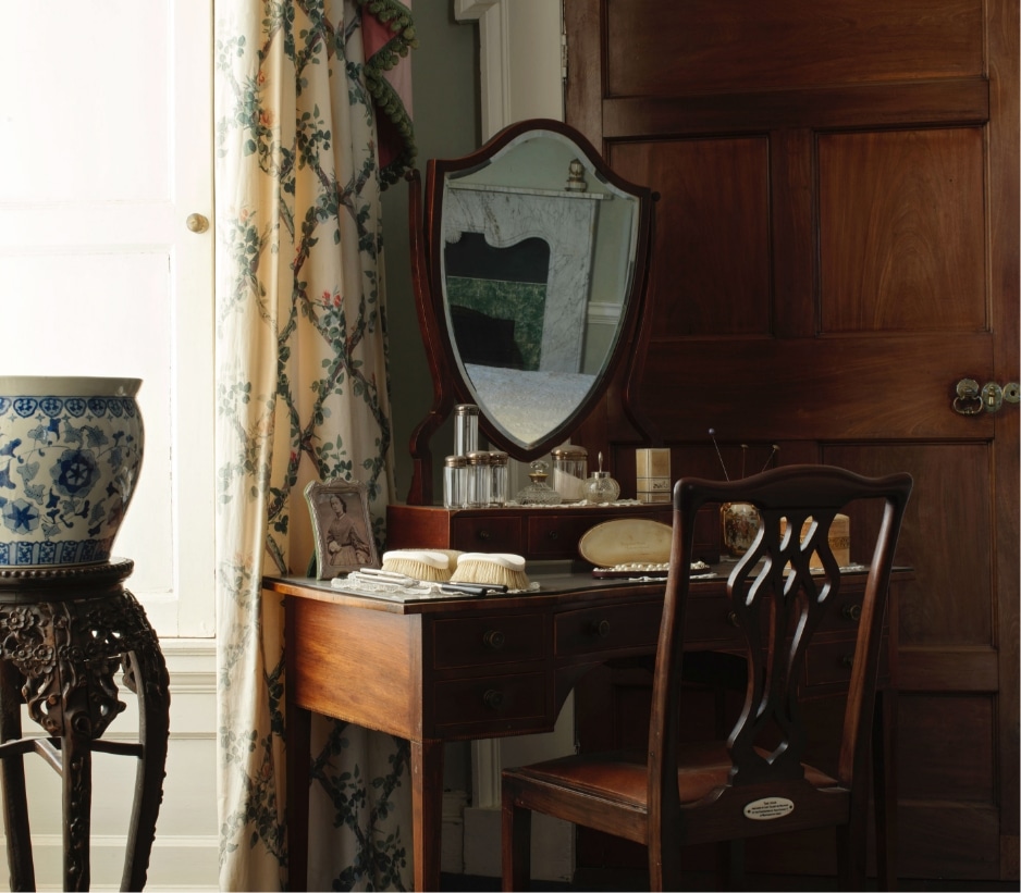 A vintage wooden vanity with an ornate mirror is set against a wooden door. It holds various items, such as brushes, perfume bottles, and a framed photograph. A blue and white ceramic vase sits on a carved stand beside floral curtains.