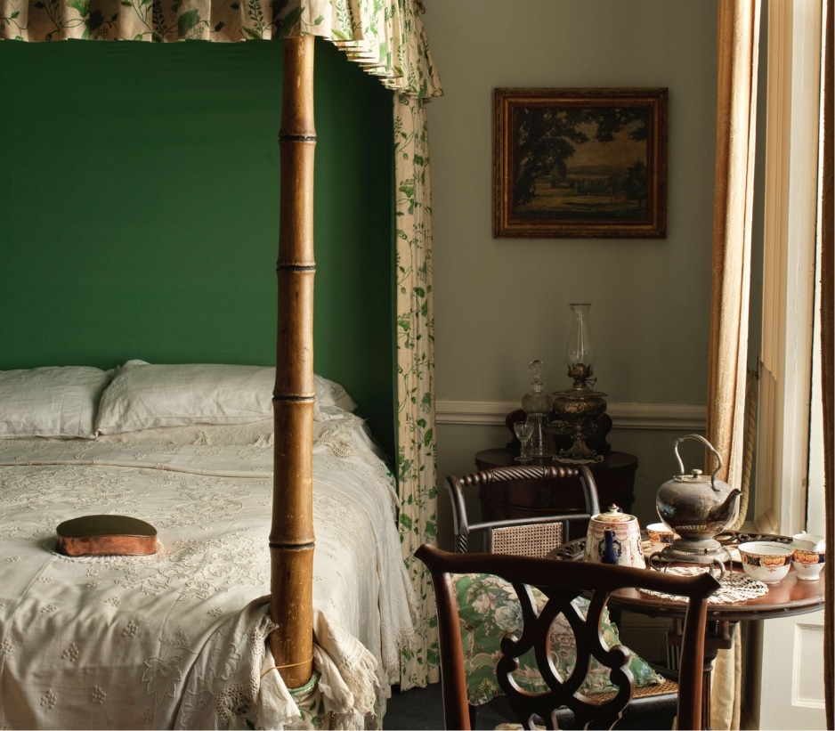 A vintage bedroom with a four-poster bed draped in floral fabric. A small table holds a teapot, cups, and plates. A framed painting hangs on the wall, and natural light streams in through a window with sheer curtains.