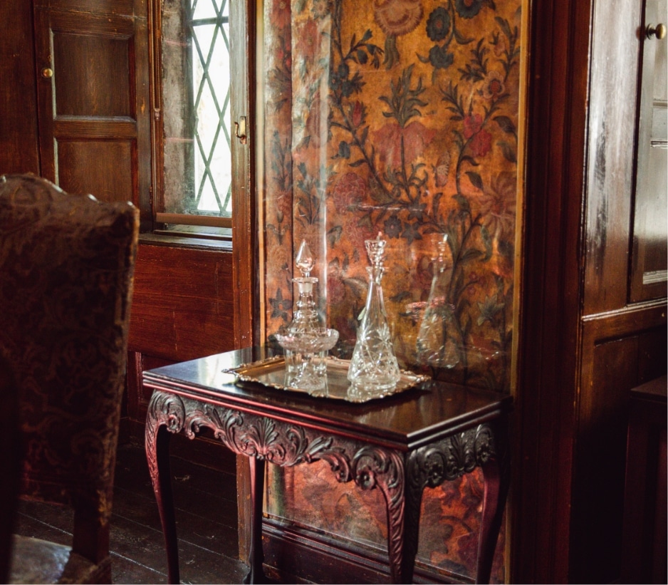 A richly detailed antique room features a wooden table with intricate carvings. On the table sits a silver tray holding an assortment of crystal decanters. The backdrop is a tapestry with floral designs, and a window with leaded glass panels.