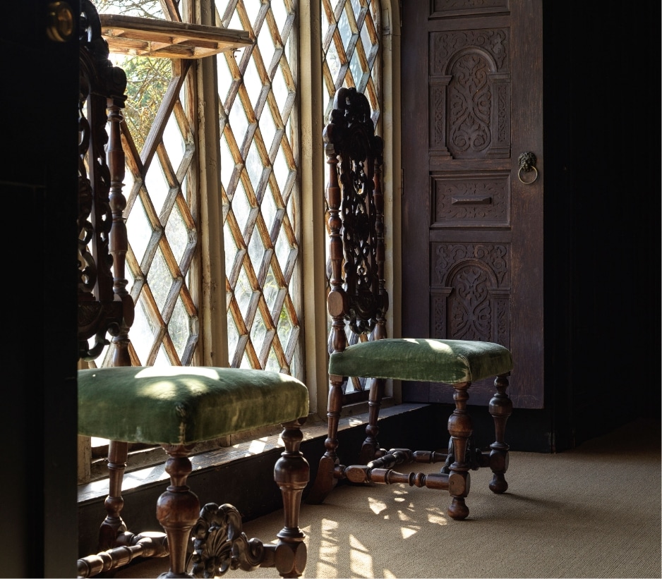 Two ornate wooden chairs with green velvet cushions sit by a large, diamond-patterned window, casting light across a dark, intricately carved wooden door and the carpeted floor.