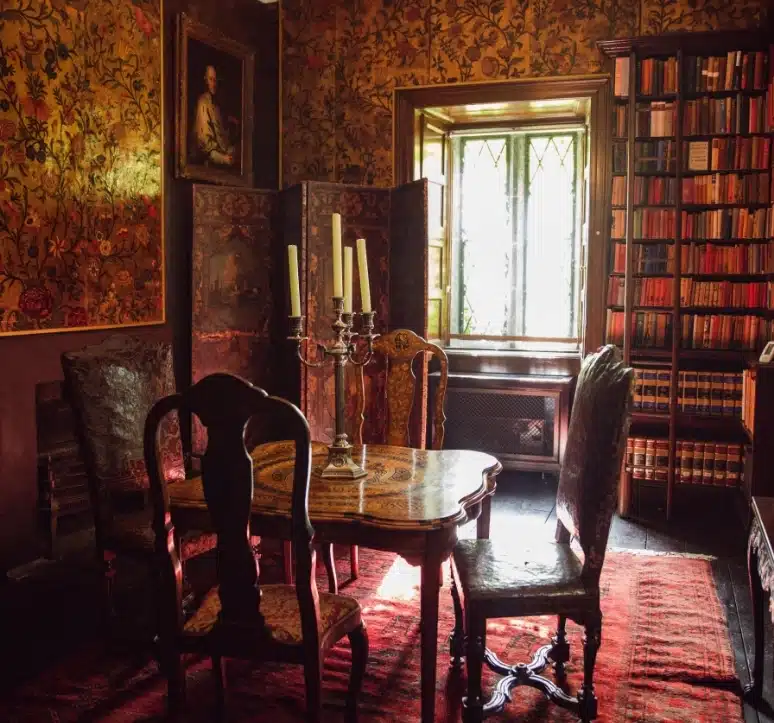 A vintage-style room with a wooden table and chairs. A candelabra sits on the table. The walls are adorned with floral wallpaper and paintings. A tall bookshelf, a patterned rug, and a window with a decorative screen complete the setting.