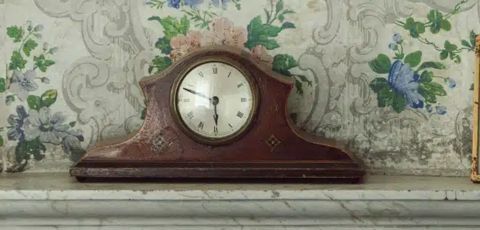 A vintage mantel clock with a wooden case sits on a marble shelf, reminiscent of the elegance found at Malahide Castle. The clock face displays Roman numerals set against a backdrop of floral-patterned wallpaper in soft greens, blues, and pinks.