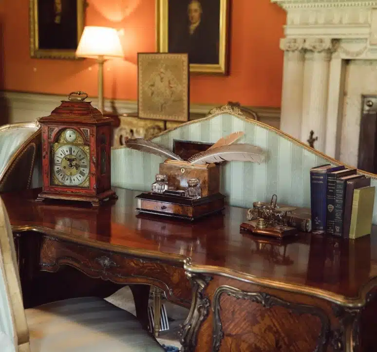 An ornate antique desk with a large clock, a quill in an inkwell, decorative books, and vintage items. A warm lamp illuminates the room, with portraits hanging on the peach-colored walls and a white fireplace in the background.