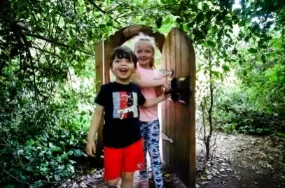 Two children stand in front of a wooden gate surrounded by lush greenery at Malahide Castle. One wears a black shirt and red shorts, and the other sports a pink shirt and patterned leggings. They are smiling, enjoying their time in the enchanting garden setting.