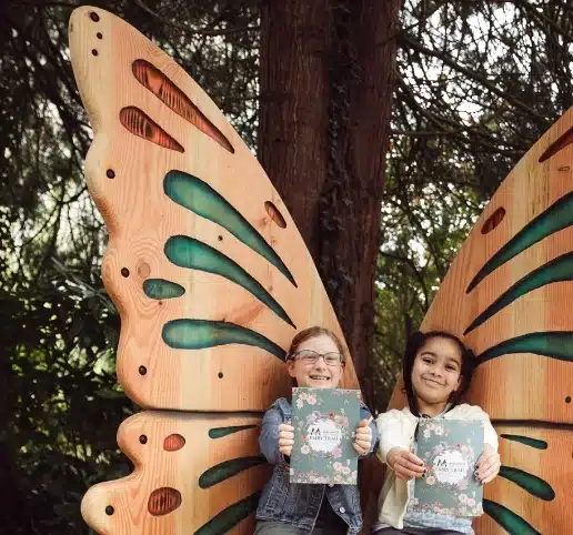 Two children sit on a wooden butterfly-shaped bench at Malahide Castle, smiling and holding books. The outdoor setting, framed by trees, creates a whimsical and natural scene.