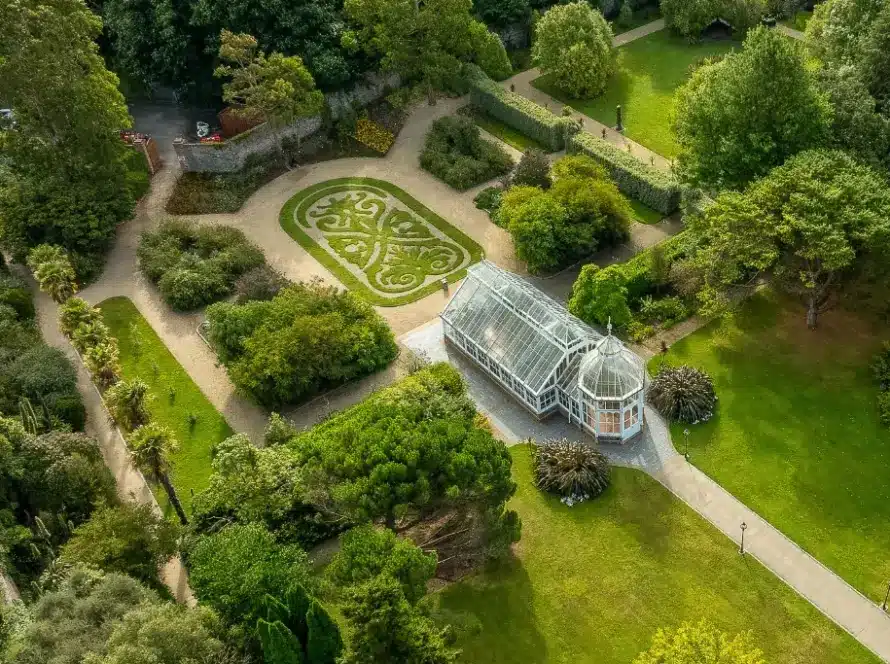 An aerial view of a botanical garden featuring a greenhouse with a glass roof and intricate design. Surrounding paths and greenery include manicured lawns, various trees, and bushes. A carefully tended ornamental garden with symmetrical patterns is visible nearby.