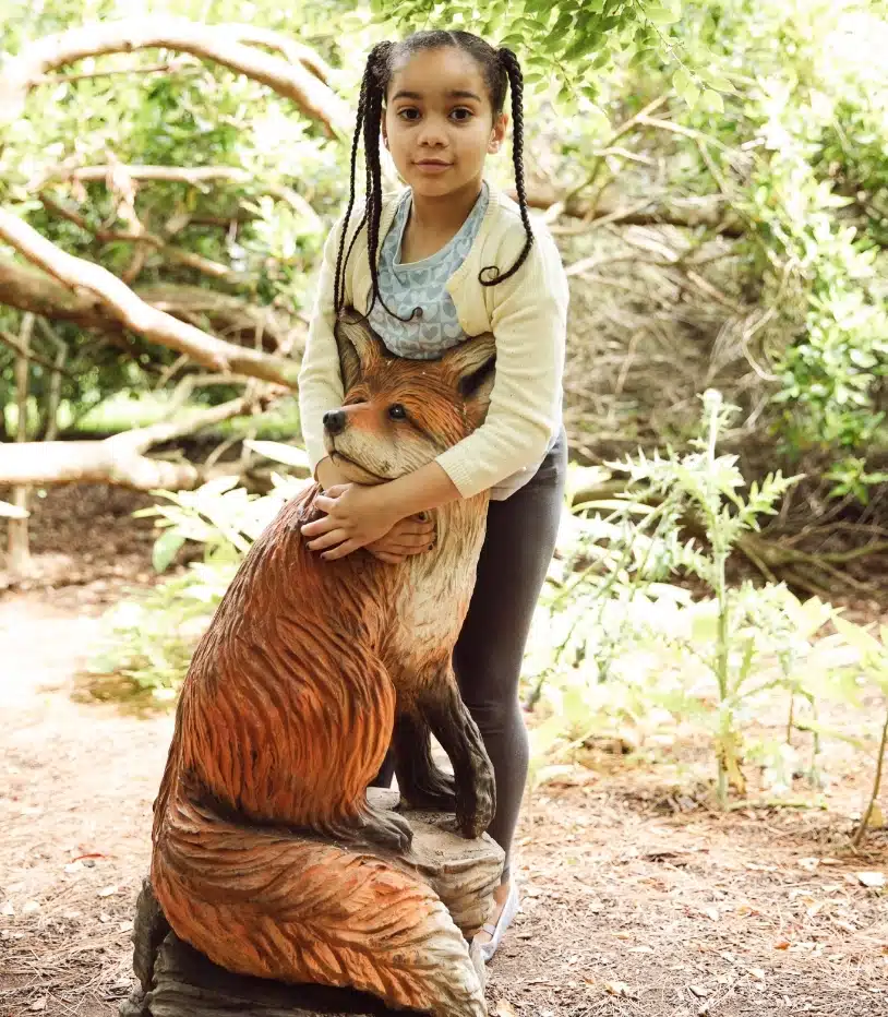 A child with braided hair is standing outdoors, embracing a large wooden fox sculpture. The backdrop reveals the lush greenery and wooded area of Malahide Castle's enchanting grounds.