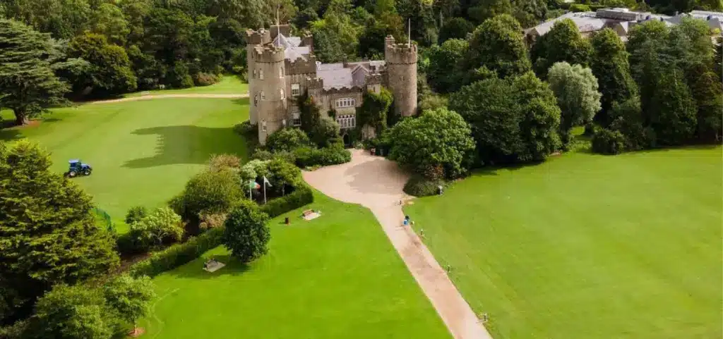 Aerial view of Malahide Castle surrounded by lush greenery and trees. The castle boasts multiple towers and an expansive lawn in front, dotted with pathways and scattered benches. A tractor is visible on the left side of the lawn, adding a touch of rustic charm.