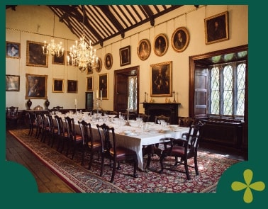 A grand dining room reminiscent of Malahide Castle features a long table set for a meal, surrounded by wooden chairs. Framed portraits adorn the walls, and elegant chandeliers hang from the ceiling. Sunlight streams through large, patterned windows onto a richly patterned rug below.