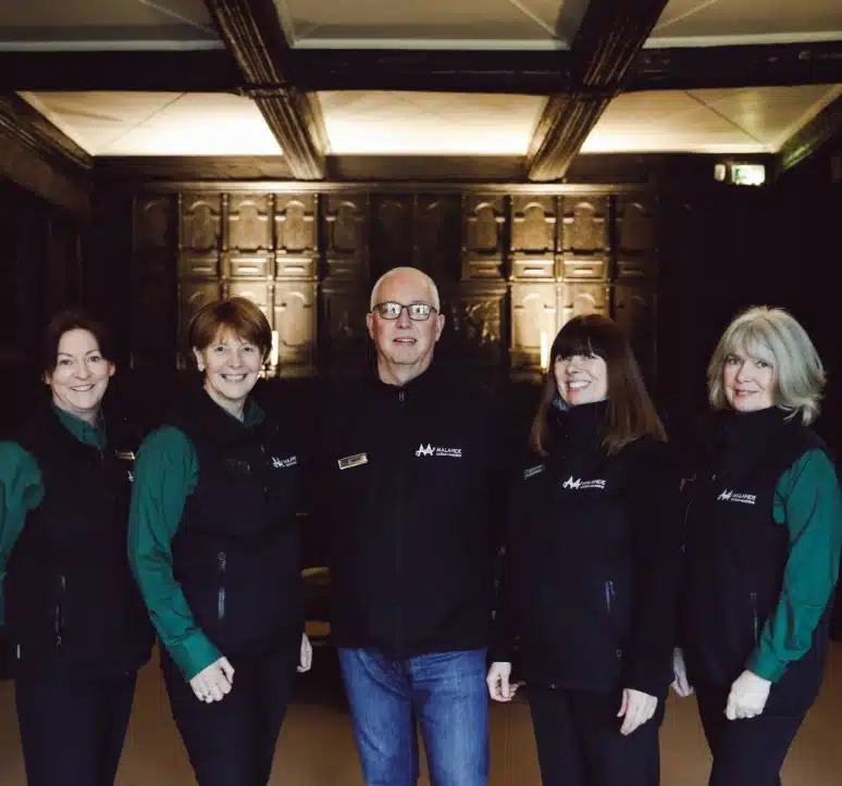 Five people stand together indoors, wearing matching dark vests and green shirts. They are smiling, and the background features dark wood paneled walls.