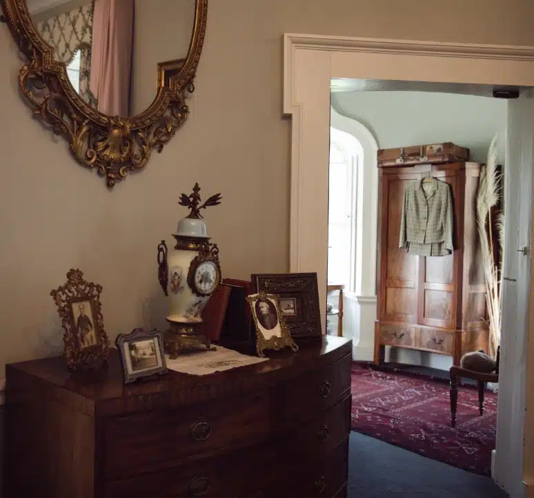 A vintage-style room featuring a wooden dresser with ornate photo frames, a decorative vase, and a gilded mirror above. An open door reveals a second room with a wooden wardrobe, a hanging coat, and a red patterned rug.