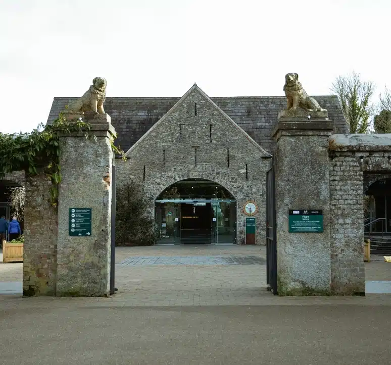 Stone entrance with two lion statues atop pillars, leading to a rustic brick building with a triangular roof and glass doors, surrounded by greenery. Informational signs are mounted on each pillar.