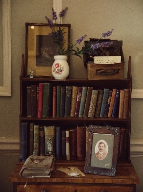 A vintage wooden bookshelf contains an assortment of old books, evoking the charm of Malahide Castle. On top, a floral vase with purple flowers, a framed portrait, and a small decorative box complement an old framed photo leaning behind the vase, adding a nostalgic touch.