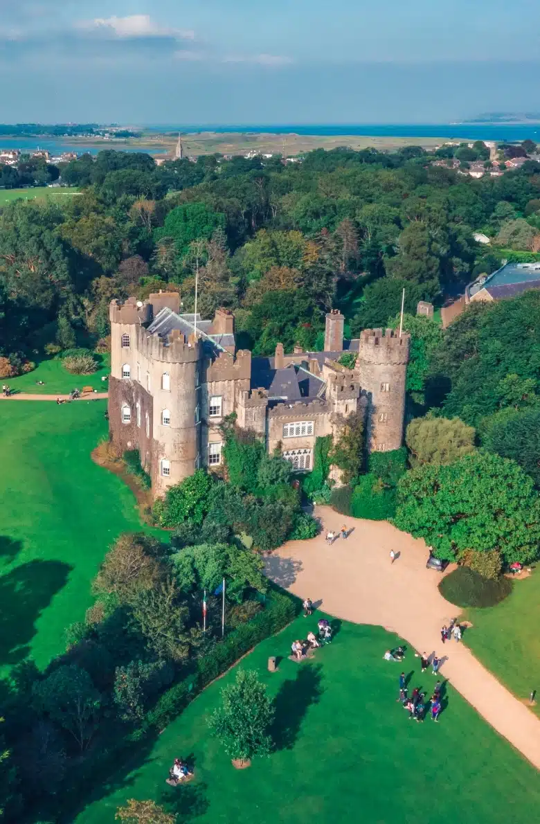 From an aerial view, Malahide Castle stands majestically, an old stone fortress encircled by lush greenery. Its multiple towers overlook a courtyard bustling with visitors. Beyond, a sea of trees stretches to the horizon where it kisses a shimmering body of water beneath a partly cloudy sky.