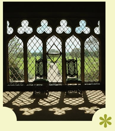 Two ornate chairs sit by a large Gothic-style window with patterned glass at Malahide Castle, casting intricate shadows on the floor. Outside, a view of the lush green landscape is beautifully visible.