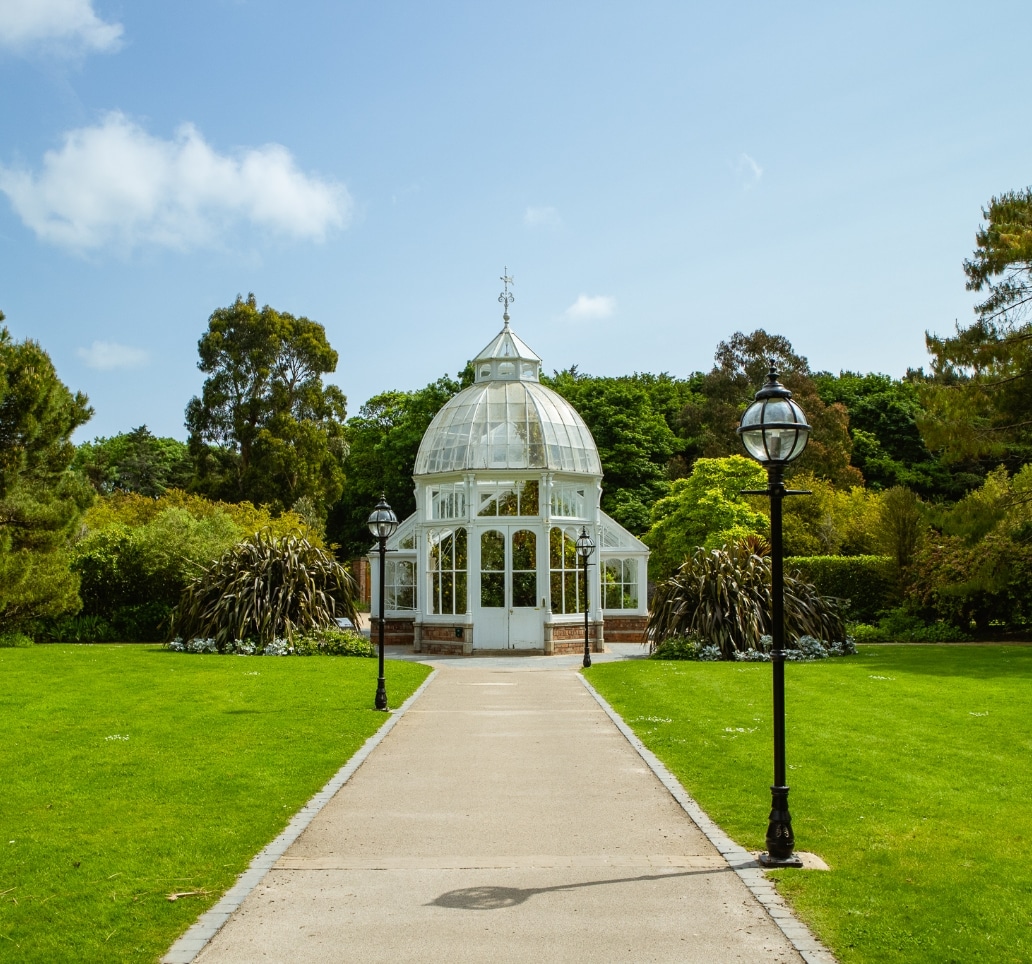 A picturesque scene featuring a white, glass-domed conservatory at the end of a paved path within the grounds of Malahide Castle. It's surrounded by lush greenery under a clear blue sky, with two vintage lampposts lining the path.