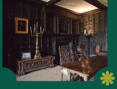 The dimly lit, historical room at Malahide Castle features dark wood paneling, an ornate carved fireplace, a large wooden table with high-backed chairs, and an intricate candelabrum. The atmosphere remains quintessentially vintage and elegant.