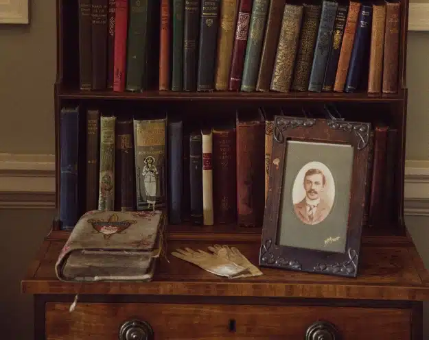 A wooden bookshelf holds an assortment of vintage books, reminiscent of those found in Malahide Castle. In front, a framed sepia-toned portrait and a pair of cream gloves rest on a wooden desk, alongside a closed, worn book with a butterfly design. The setting is warm and nostalgic.