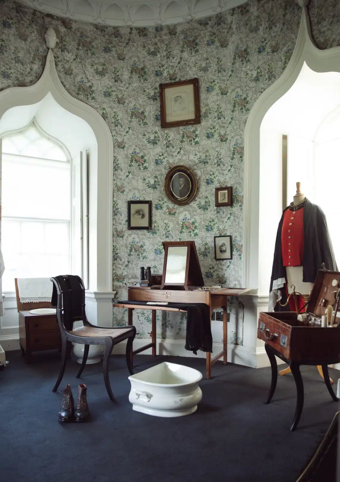 A beautifully decorated vintage room with ornate wallpaper and arched windows offers a glimpse into the elegance of Malahide Castle. It features an antique vanity with a mirror, a small chair, and framed pictures on the walls. A mannequin dressed in a red vest stands next to a wooden case with grooming tools.