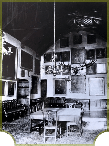 A vintage black and white photo of a grand interior room featuring a central dining table with chairs, ornate chandeliers, and walls adorned with numerous framed artworks and photographs.