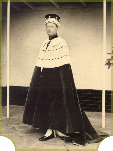 A person wearing a royal cape and crown stands on a concrete surface. The cape is long and dark with a white, fur-trimmed collar dotted with black spots. The background features a white brick wall.