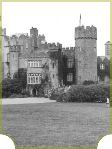 Black and white image of a large, historic castle with ivy-covered stone walls, a central tall tower, and a large arched window. The foreground features a grassy lawn with a pathway leading to the entrance.