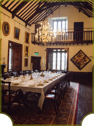 A long, elegant dining table set for a formal dinner in a grand room with wooden floors and a high ceiling. Chandeliers hang overhead, and the walls are adorned with portraits and framed artwork. A staircase leads to a balcony.