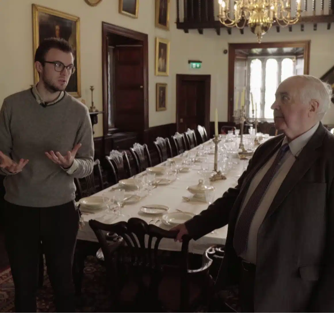 Two men stand in the historic dining room of Malahide Castle, where a long table is set for a meal. One man gestures as he speaks, while the other listens. The room features a chandelier, portraits on the walls, and a large window in the background.