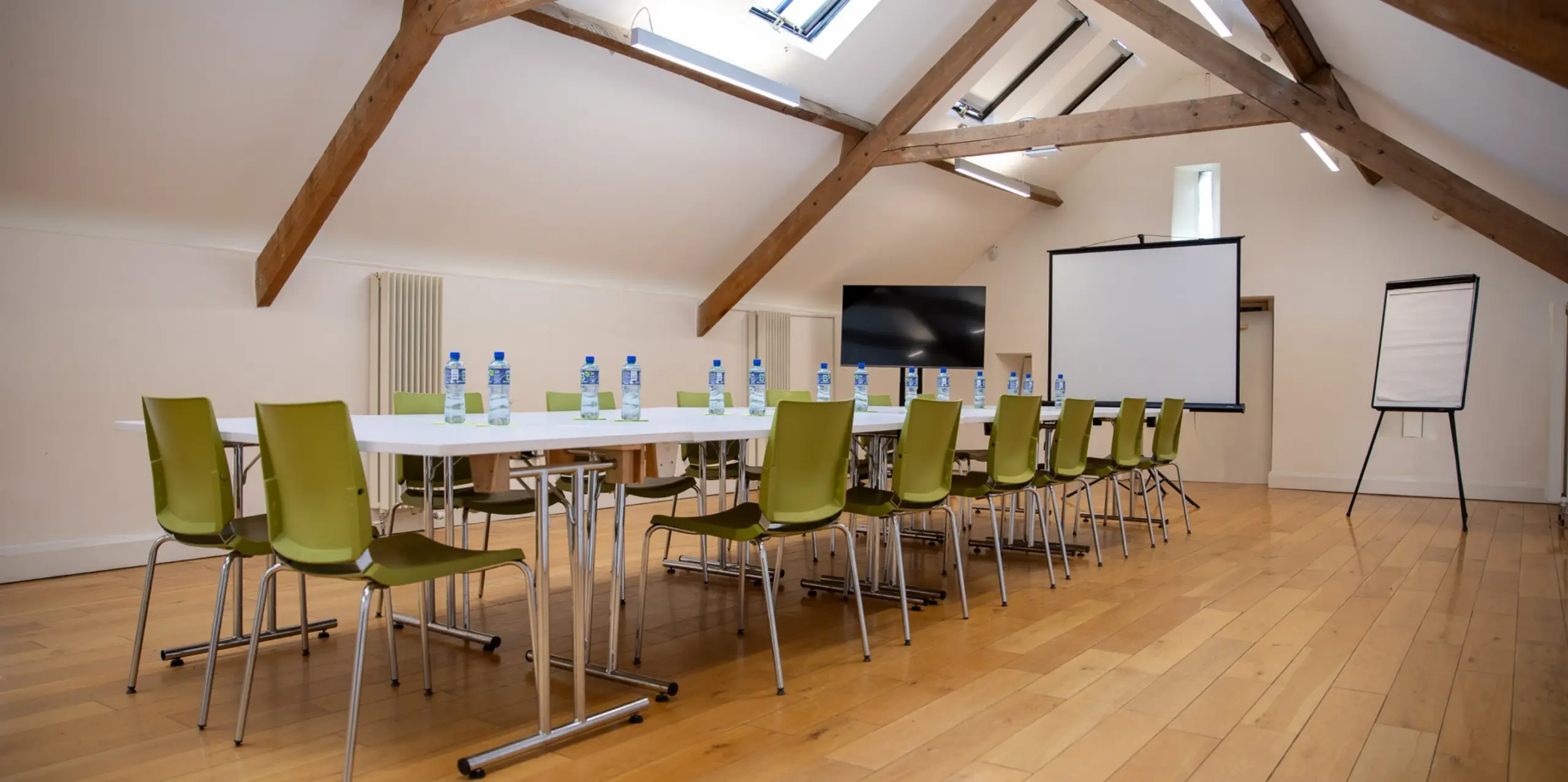 A conference room with a long table set for a meeting. Green chairs are arranged around the table, which has water bottles and glasses. A screen, flip chart, and TV are at the front. The room has wooden floors and exposed beams on the ceiling.