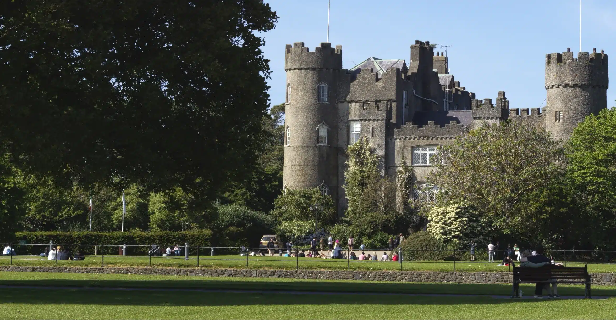 Malahide Castle, a historic fortress with stone towers and battlements, is surrounded by lush green trees and a sprawling grassy lawn. On this sunny day, visitors are sitting and strolling in the foreground, soaking in the serene ambiance of this enchanting site.