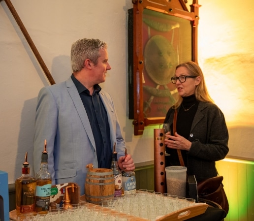 A man and woman are chatting next to a table filled with various bottles and shot glasses. The man, in a blue suit, and the woman, donning a dark coat with glasses, stand before a wooden frame adorned with details reminiscent of Malahide Castle's elegance.