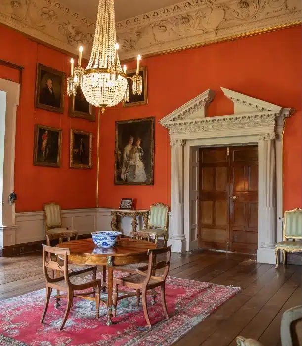 In an elegant room reminiscent of Malahide Castle, orange walls are adorned with portraits and a chandelier. A round wooden table with a decorative bowl rests on a red rug, surrounded by ornate chairs. A door with a detailed white frame graces one wall.