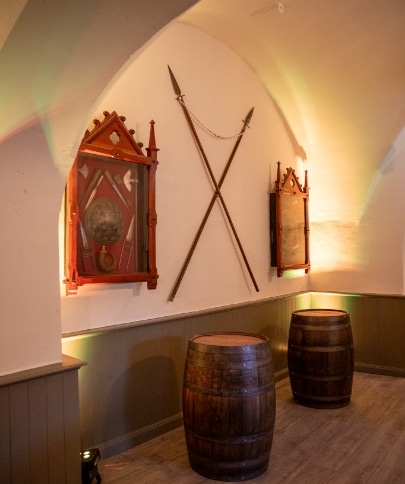 A dimly lit room with two wooden barrels on the floor evokes the rustic ambiance of Malahide Castle. Two medieval shields and crossed spears are mounted on the arched white wall, enhancing its historical charm.