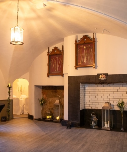 A dimly lit room in Malahide Castle features two ornate display cases on the wall containing swords. Below them, a fireplace is adorned with lanterns. A curved archway reveals a glimpse of a table set with candles in the background, enhancing the historic charm.