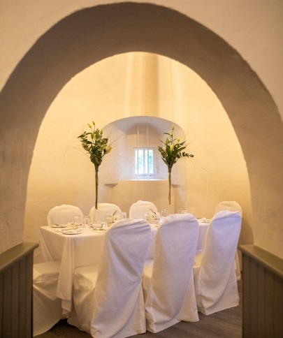 A cozy dining room reminiscent of Malahide Castle features a long table set for six, draped in white. Tall green floral arrangements grace the table, perfectly illuminated under a softly lit arched ceiling by the window.