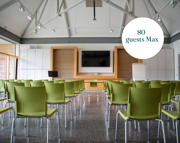 A conference room with rows of green chairs facing a wooden presentation area, reminiscent of Malahide Castle's elegant charm. A large TV and flip chart stand ready for presentations. A circular overlay reads "80 guests Max." The space boasts a modern design with exposed beams and lighting.