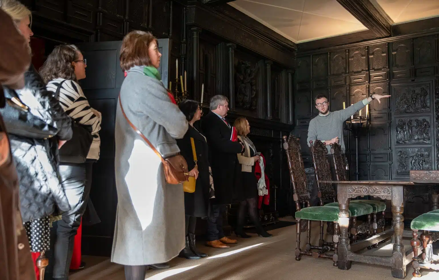 A tour guide gestures while speaking to a group inside a historic room at Malahide Castle, where dark wood paneling and ornate chairs add to the grandeur. The attendees stand in a semi-circle, attentively listening.