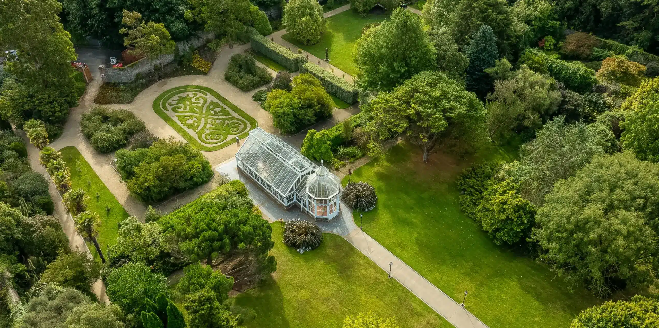 Aerial view of a botanical garden with a prominent glass conservatory in the center. Surrounding paths, manicured lawns, and intricately designed hedges enhance the lush greenery and diverse trees.