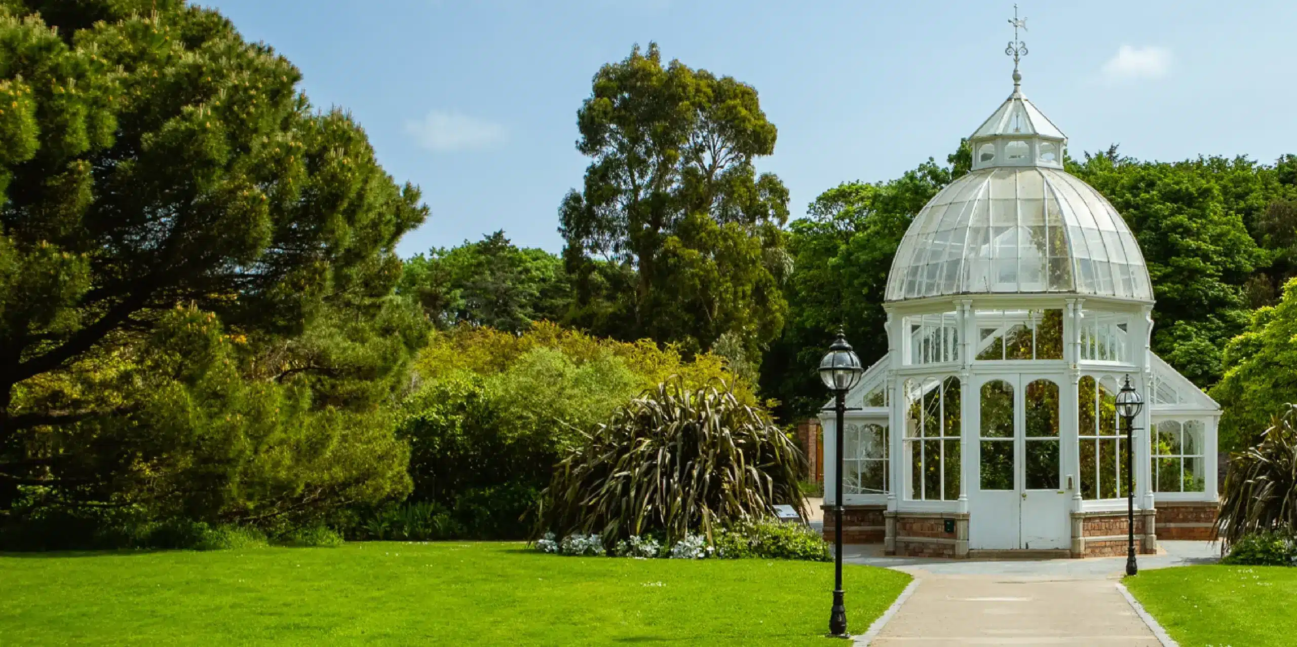 A small, white, glass conservatory is surrounded by lush green trees and a well-maintained lawn. A pathway leads to its entrance, and an ornate lamp post stands nearby. The sky is clear and blue, indicating a sunny day.