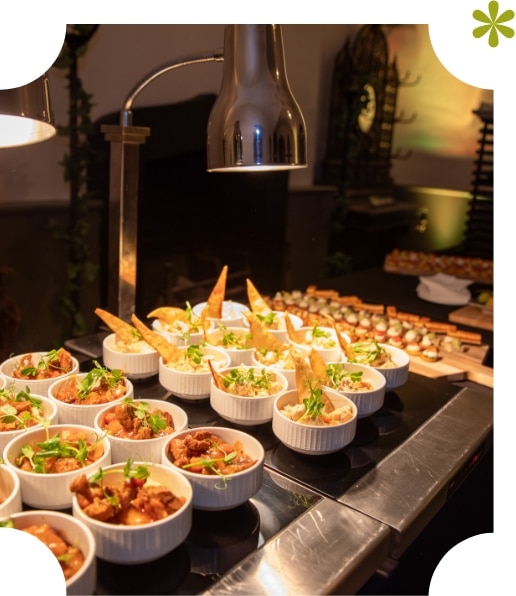 A buffet table at Malahide Castle features small white bowls of savory dishes garnished with greens and crispy chips, warmed by a lamp. In the background, an array of desserts and pastries is elegantly displayed on a wooden board in the dimly lit setting.