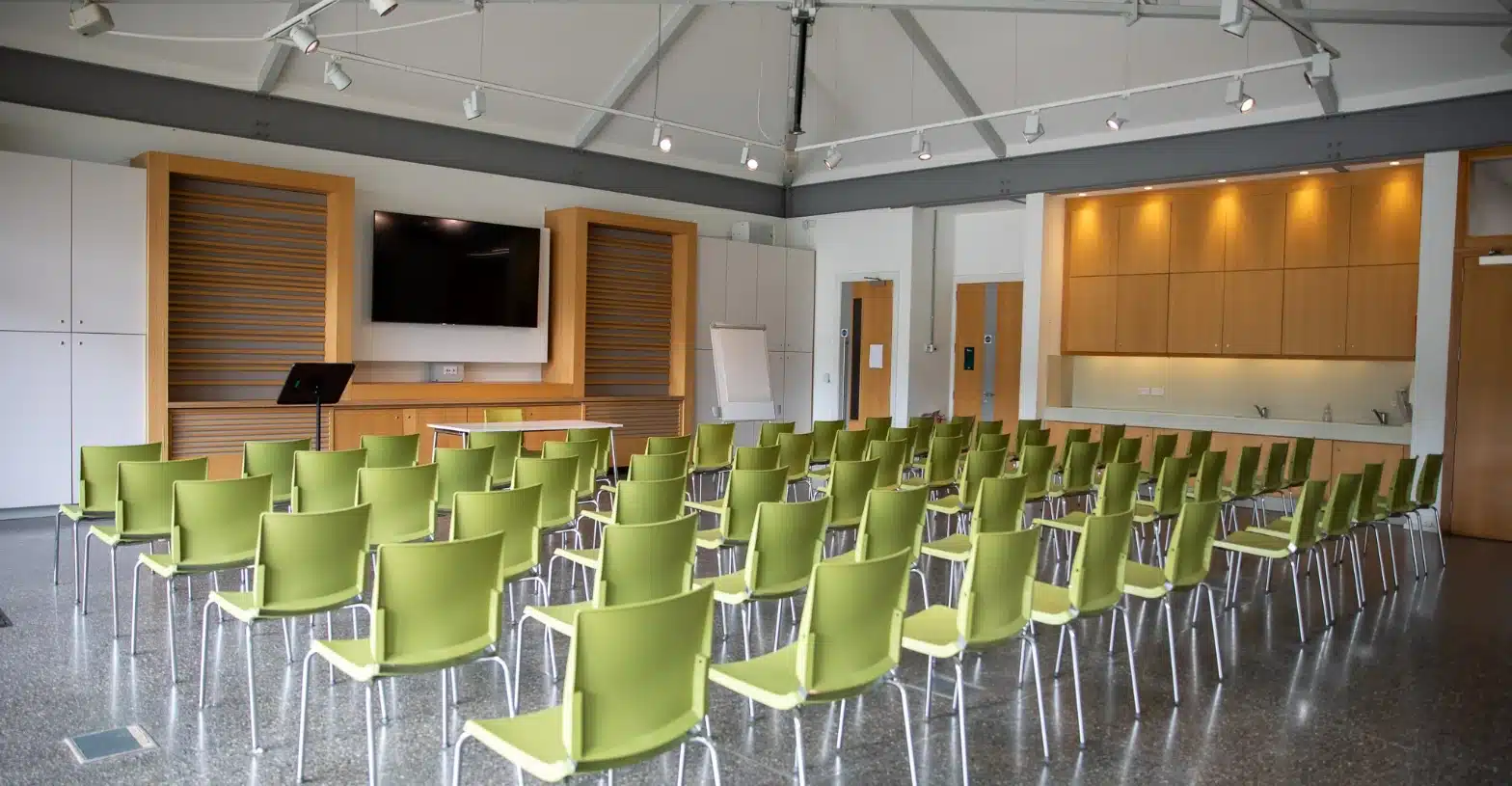 A modern conference room with green chairs in rows facing a large flat-screen TV exudes a touch of Malahide Castle elegance. Wooden paneling, a podium, and a whiteboard enhance the space, while high ceilings with track lighting and a kitchenette on the right complete the stylish setup.