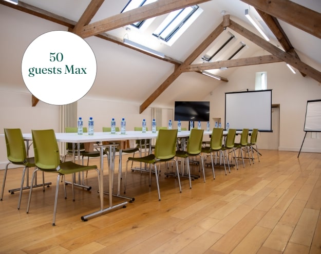 A conference room with wooden floors and exposed beams exudes the charm of Malahide Castle. It features two long tables with water bottles and green chairs. At the back, a projector screen and flip chart stand ready. A sign reads "50 guests Max.