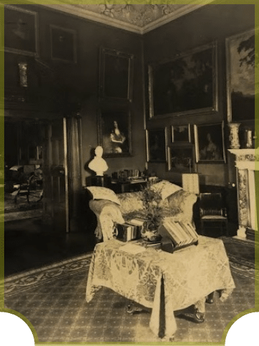 A vintage room with ornate furniture, featuring a central table covered with a cloth and books, surrounded by chairs. The walls are adorned with framed paintings and a bust, next to a fireplace. The decor suggests a classical, elegant style.