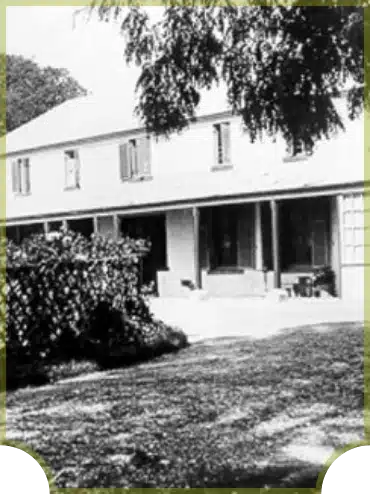 A black and white photo of a large, two-story house with a covered porch, surrounded by trees and bushes.