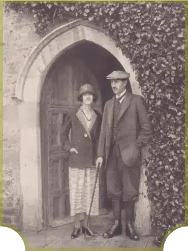 A vintage photo of a woman in a long coat and hat standing beside a man in a suit with a cap and cane. They are in front of an arched wooden door with ivy growing on the stone wall.